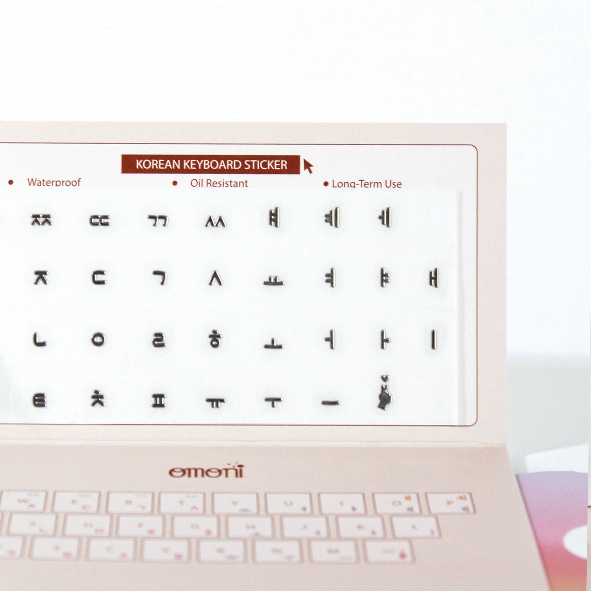 A zoomed detail image showing a laptop keyboard with Korean stickers applied to it, alongside a package with a sticker design visible through the seal. Seamless die-cut Korean Hangul keyboard sticker with Korean alphabet letters for learning Korean, typing practice, vocabulary building and beginner Korean language lessons. Metallic Dark Gray.