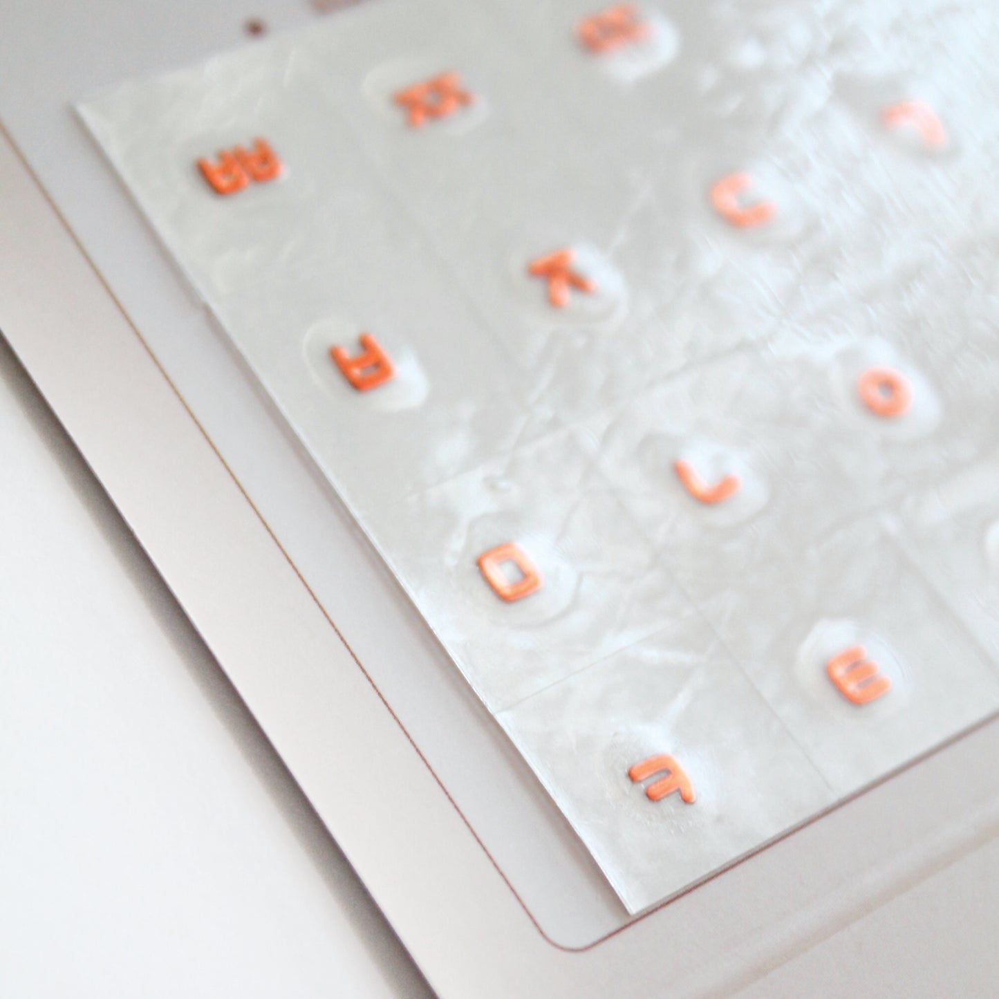 A zoomed detail image showing a laptop keyboard with Korean stickers applied to it, alongside a package with a sticker design visible through the seal. Seamless die-cut Korean Hangul keyboard sticker with Korean alphabet letters for learning Korean, typing practice, vocabulary building and beginner Korean language lessons. Metallic Citrus.