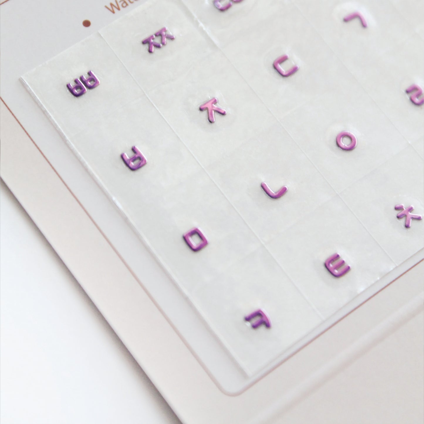 A zoomed detail image showing a laptop keyboard with Korean stickers applied to it, alongside a package with a sticker design visible through the seal. Seamless die-cut Korean Hangul keyboard sticker with Korean alphabet letters for learning Korean, typing practice, vocabulary building and beginner Korean language lessons. Metallic Violet.