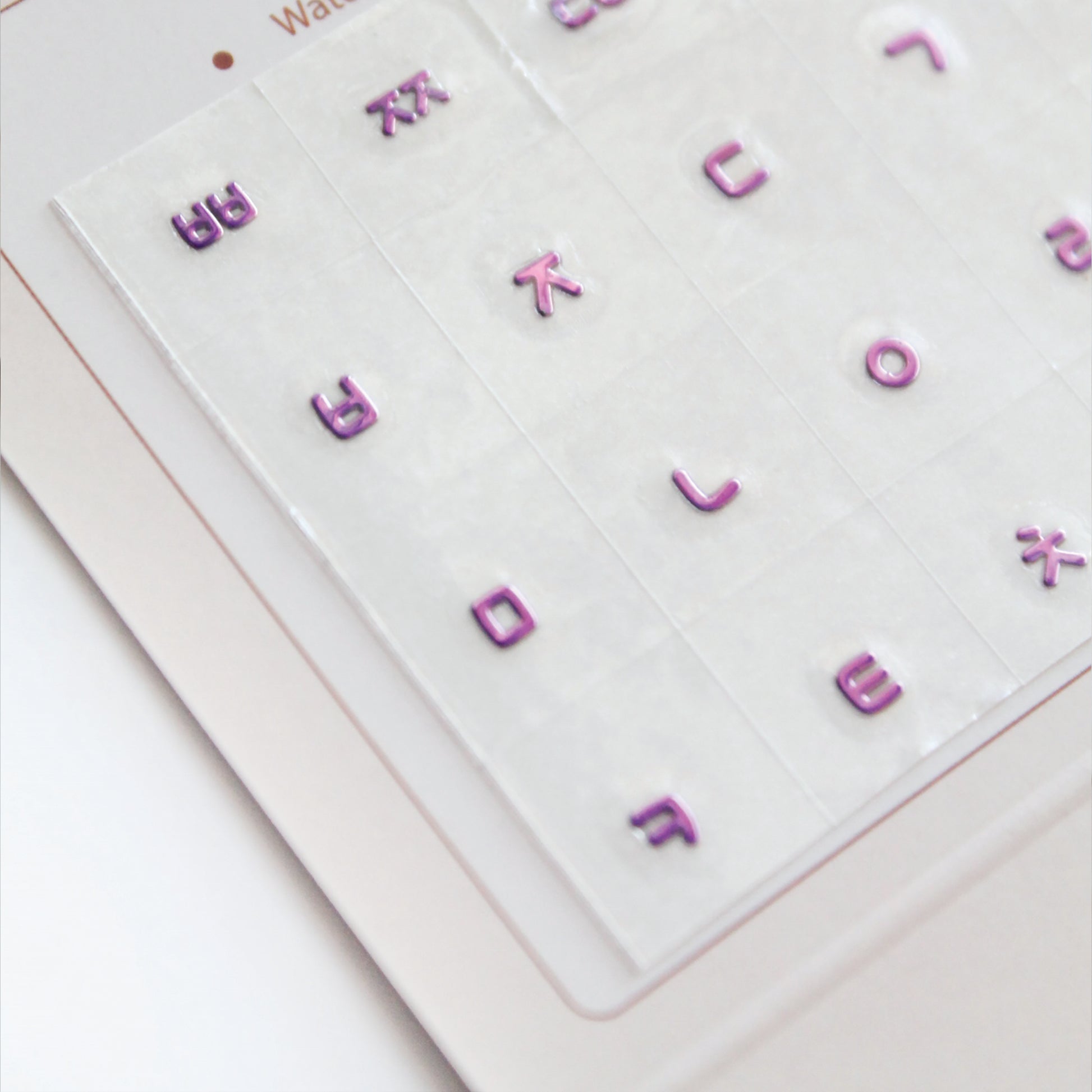 A zoomed detail image showing a laptop keyboard with Korean stickers applied to it, alongside a package with a sticker design visible through the seal. Seamless die-cut Korean Hangul keyboard sticker with Korean alphabet letters for learning Korean, typing practice, vocabulary building and beginner Korean language lessons. Metallic Violet.